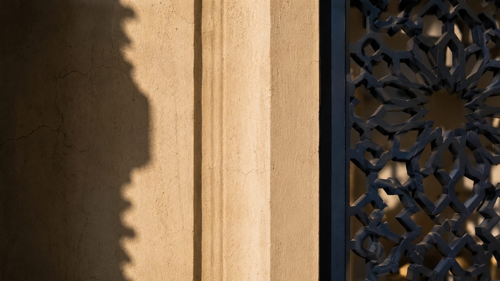 Sunlight and shadow across a stone facade beside a geometric mashrabiya screen